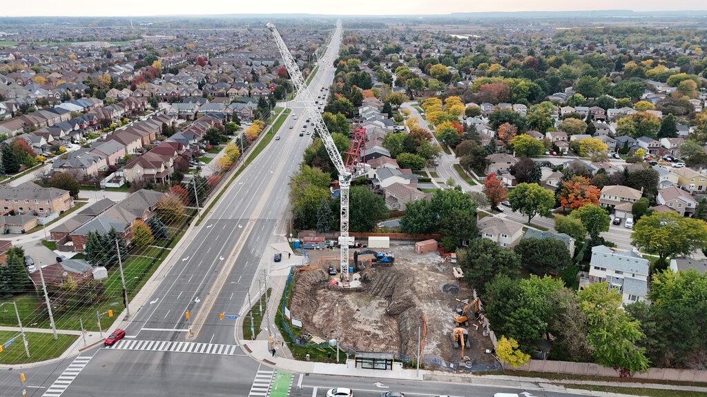 Seen from high above, a white tower crane stands over the excavated corner lot, with the boulevard running off between autumn-coloured neighbourhoods.