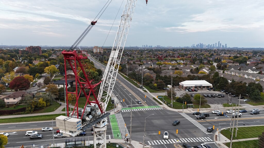 Level with the top of the red and white tower crane, looking out over a busy intersection and tree-lined streets toward a city skyline on the horizon.