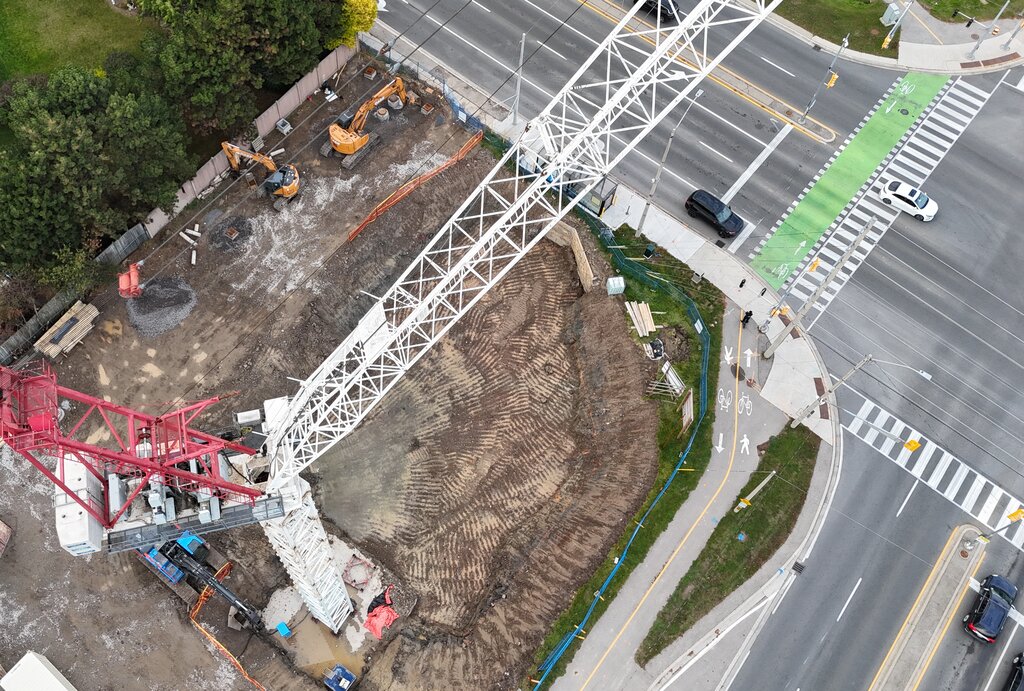 Directly overhead, the crane's red and white arms reach across the excavation, with excavators along the back fence and a painted bike crossing at the corner.