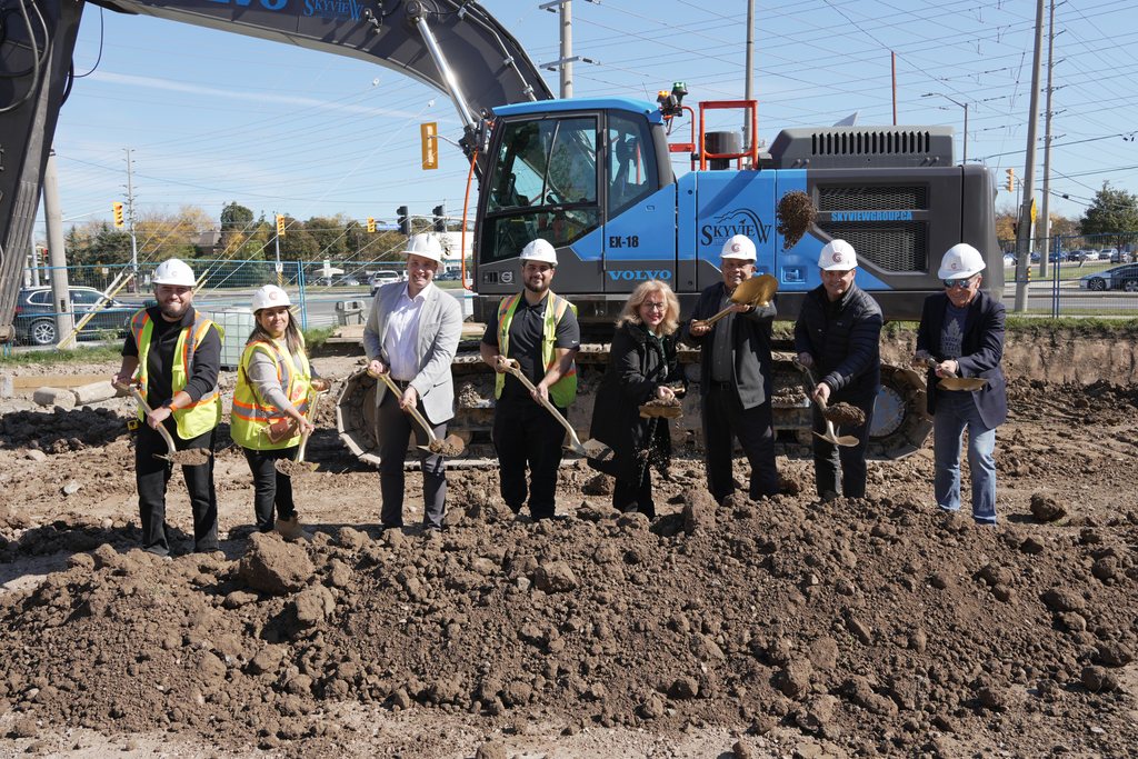 A line of people in white hard hats, some in safety vests, holding gold shovels in the turned earth with the excavator behind.