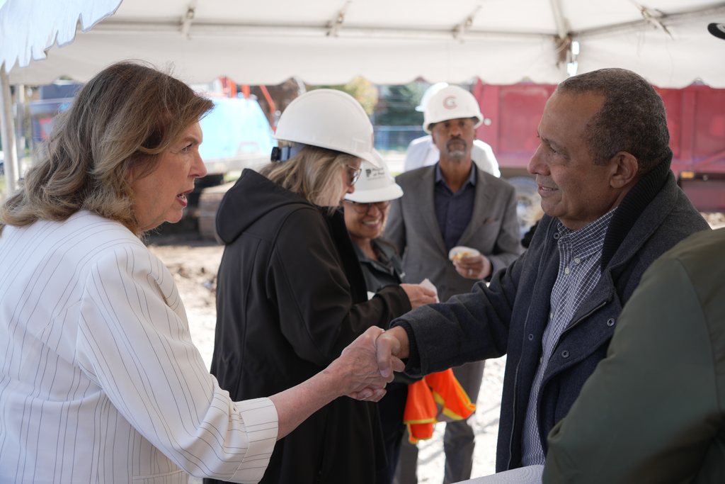 Two guests shake hands and smile under the tent while others in hard hats look on.