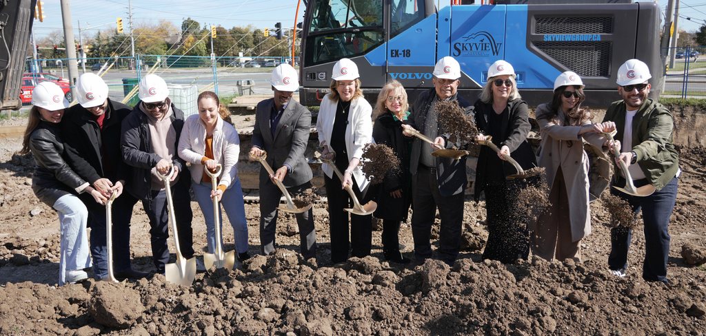 Guests in white hard hats throw the first shovels of earth together, soil in the air, in front of a blue excavator on the site.
