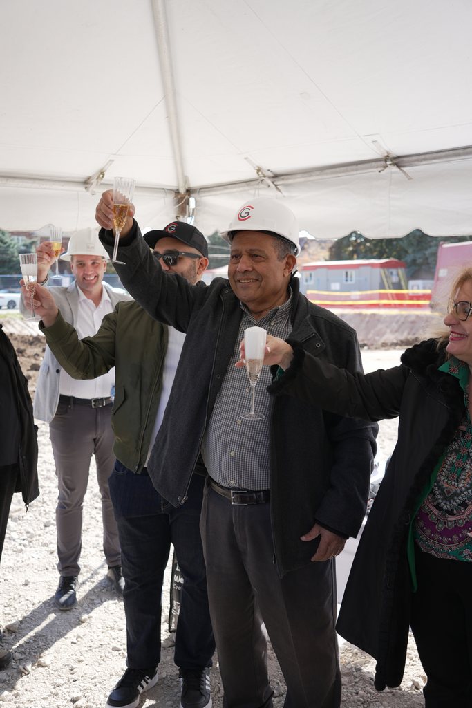 Under the tent, a man in a white hard hat raises a glass of sparkling wine with guests around him.
