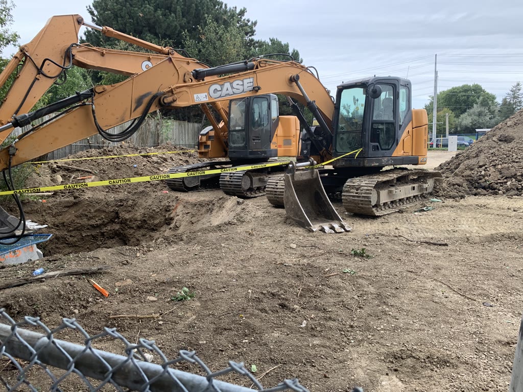 Two excavators beside a freshly dug excavation, with caution tape strung around the opening and mounds of soil.