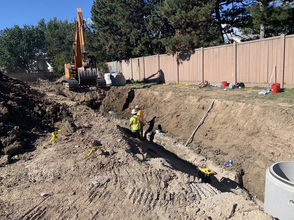 A long, deep trench runs beside the site's back fence, with an excavator at the far end and two workers in hard hats and safety vests in the trench.