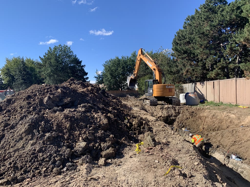 A worker in a safety vest bends over a pipe in the trench while an excavator works beside a large pile of excavated earth.