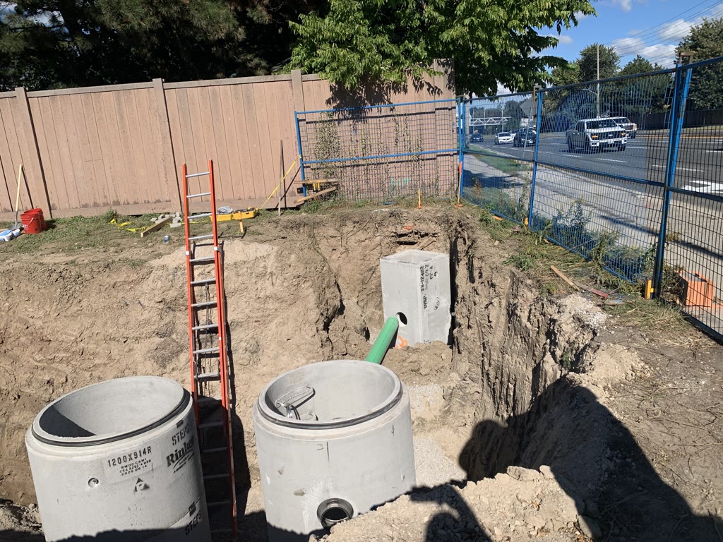 Precast concrete manhole sections and a green pipe set in a deep excavation at the edge of the site, with a ladder leading down.
