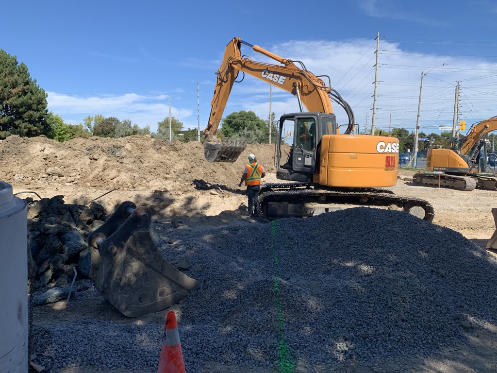 An excavator and a pile of crushed gravel on the site, with a worker in a safety vest and a second excavator behind.