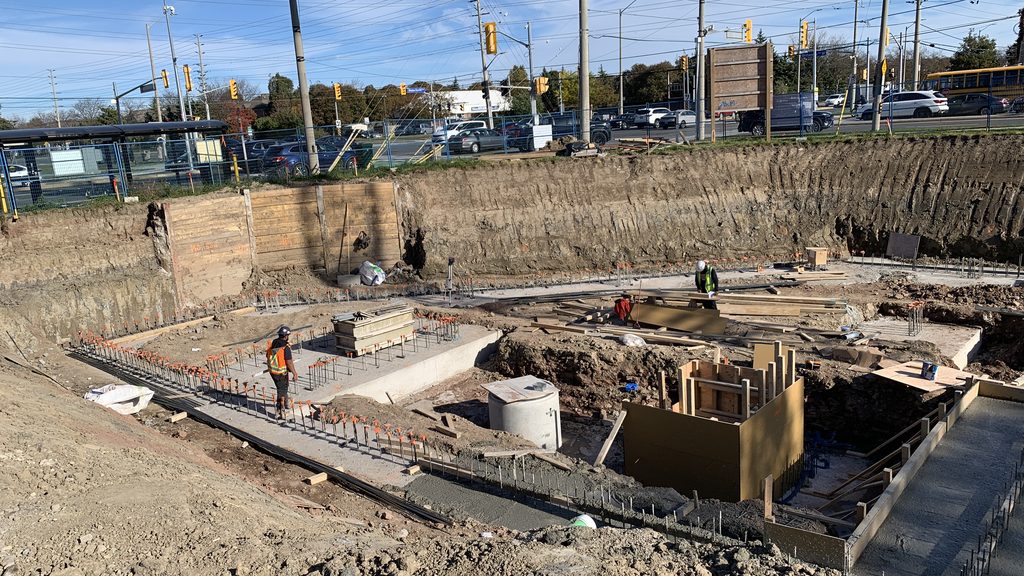 Inside the excavation, a long concrete footing with rows of steel reinforcing bars standing in it, a worker in a hard hat and safety vest walking along it, timber forms and a precast concrete ring nearby, and the boulevard beyond the shoring.