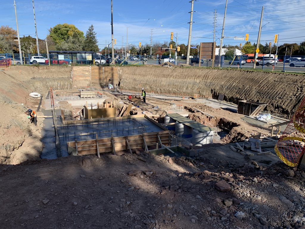 A wide view of the excavation with poured concrete pads, timber forms and precast concrete rings, two workers in safety vests, and the boulevard and a bus shelter beyond.
