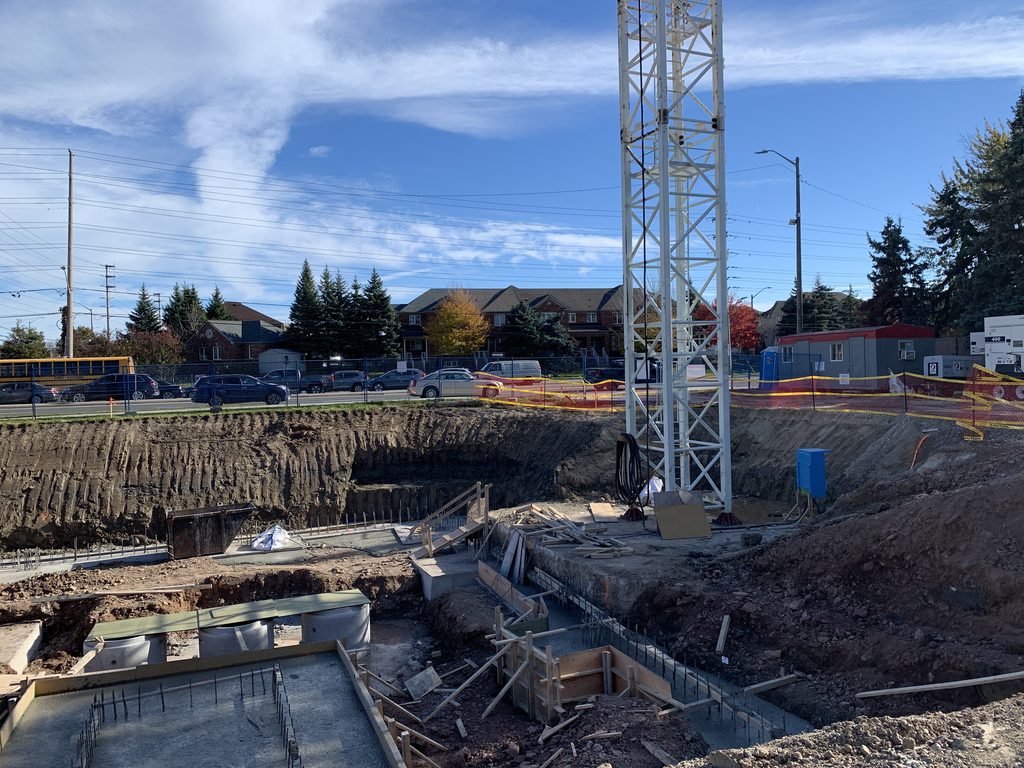 The white lattice mast of the tower crane rising from the excavation beside freshly poured footings, with site trailers beyond the safety fence.